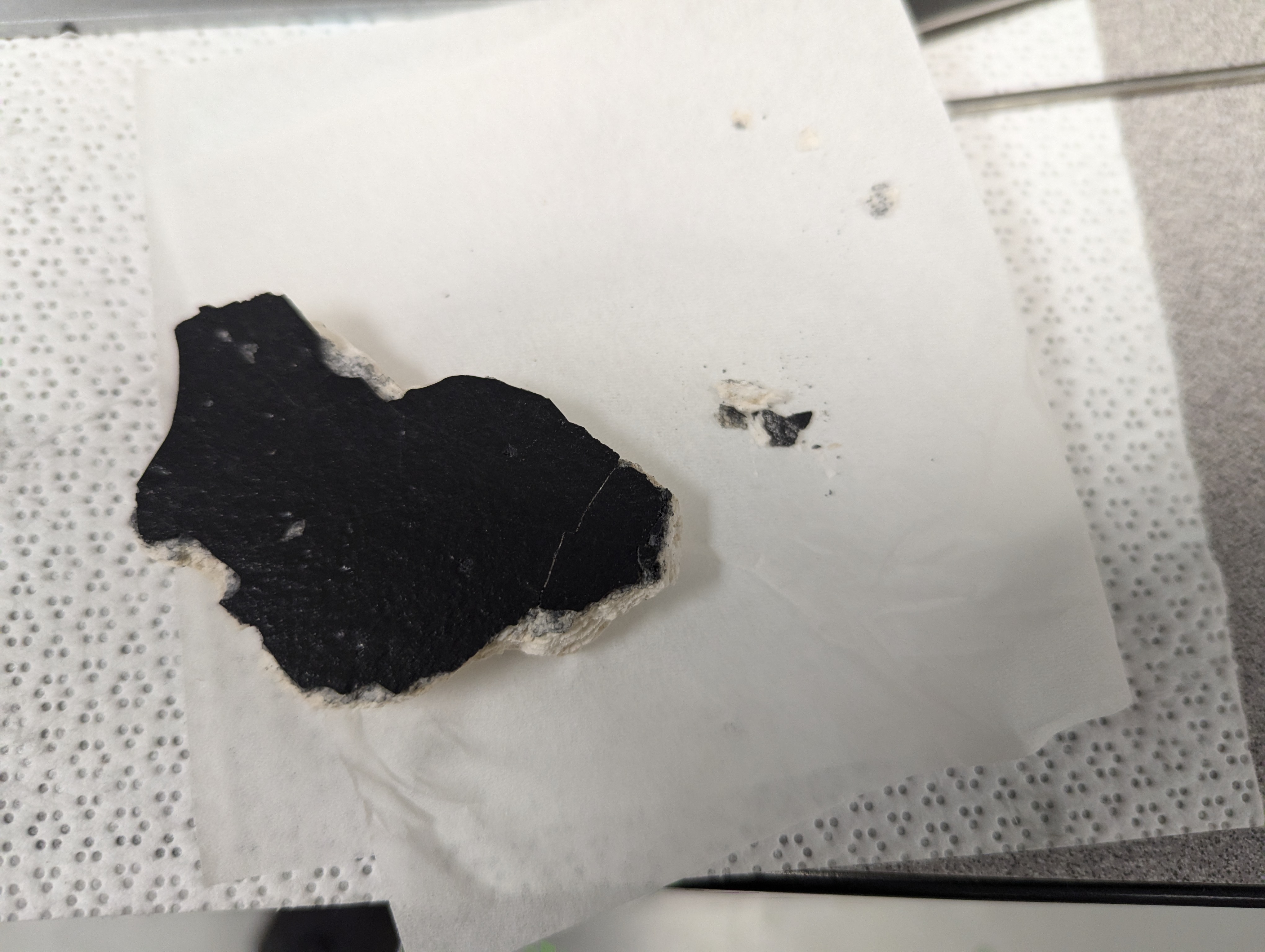 The tile fragment. The black outer coating sits on top of the white fibrous body, visible along the broken edge.